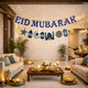 EID Mubarak Luxe Banner hanging across a modern, warmly lit living room decorated for festive Eid celebrations with candles and floral arrangements.
