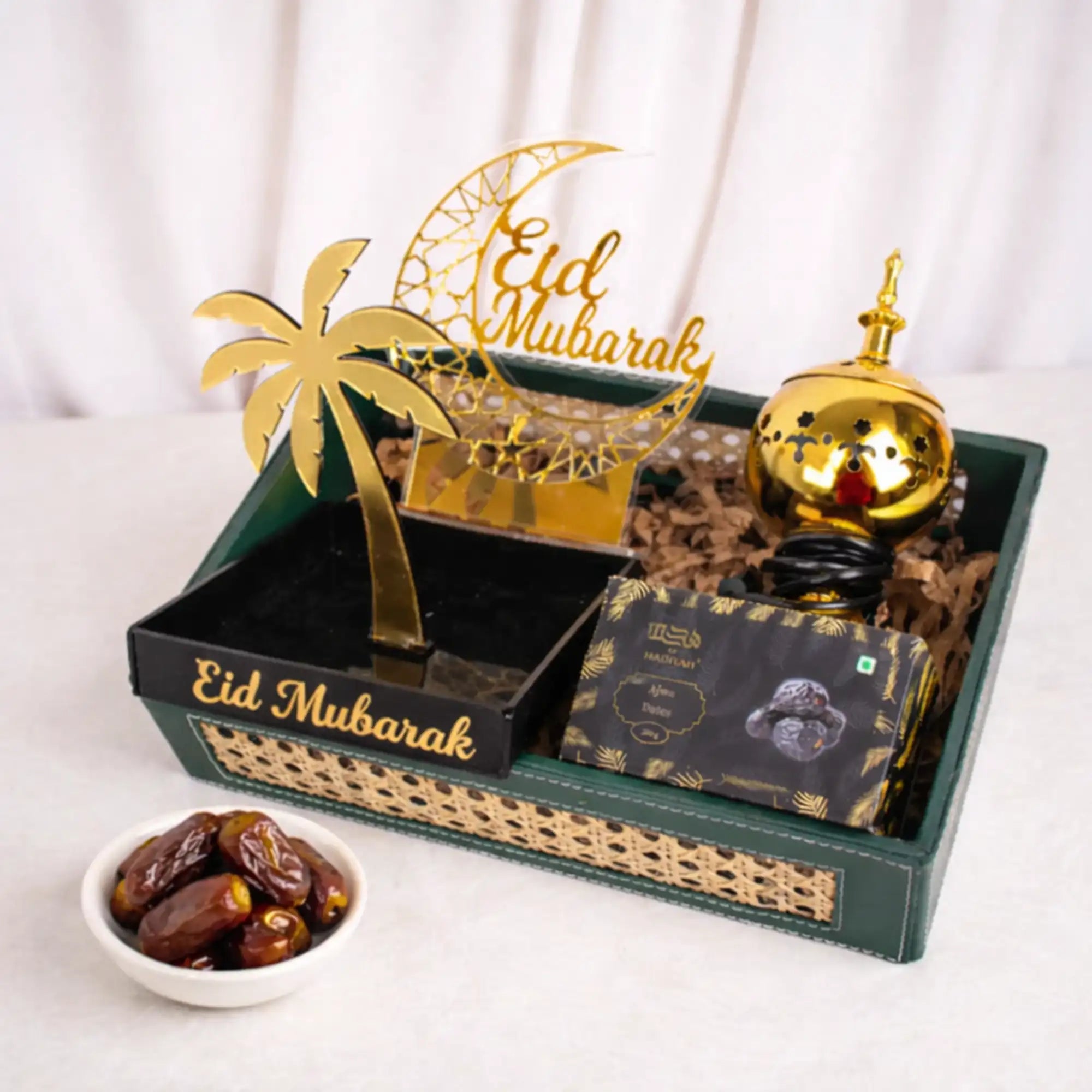 A festive Eid gift set displayed on a white surface, featuring a gold electric bakhoor burner, a gold crescent moon 'Eid Mubarak' stand, a black and gold palm tree tray, and a box of Ajwa dates with a few loose dates nearby. Pink artificial blossoms decorate the background. Main image