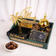 A festive Eid gift set displayed on a white surface, featuring a gold electric bakhoor burner, a gold crescent moon 'Eid Mubarak' stand, a black and gold palm tree tray, and a box of Ajwa dates with a few loose dates nearby. Pink artificial blossoms decorate the background.