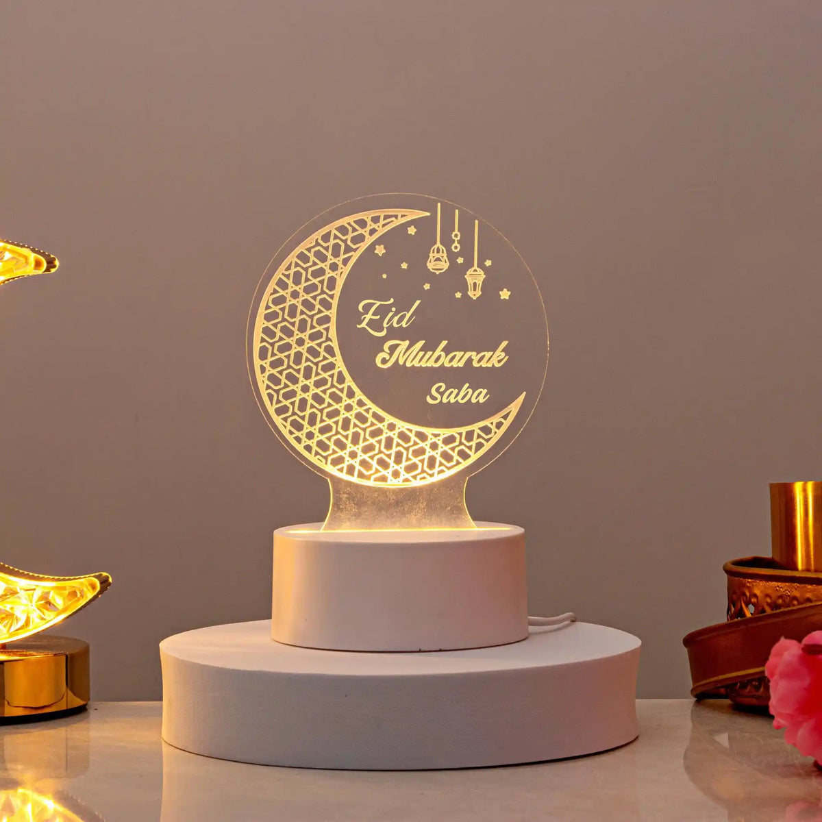Personalized Eid Mubarak LED lamp emitting a warm glow, featuring an intricate crescent moon design and the name "Saba", displayed on a white cylindrical base.