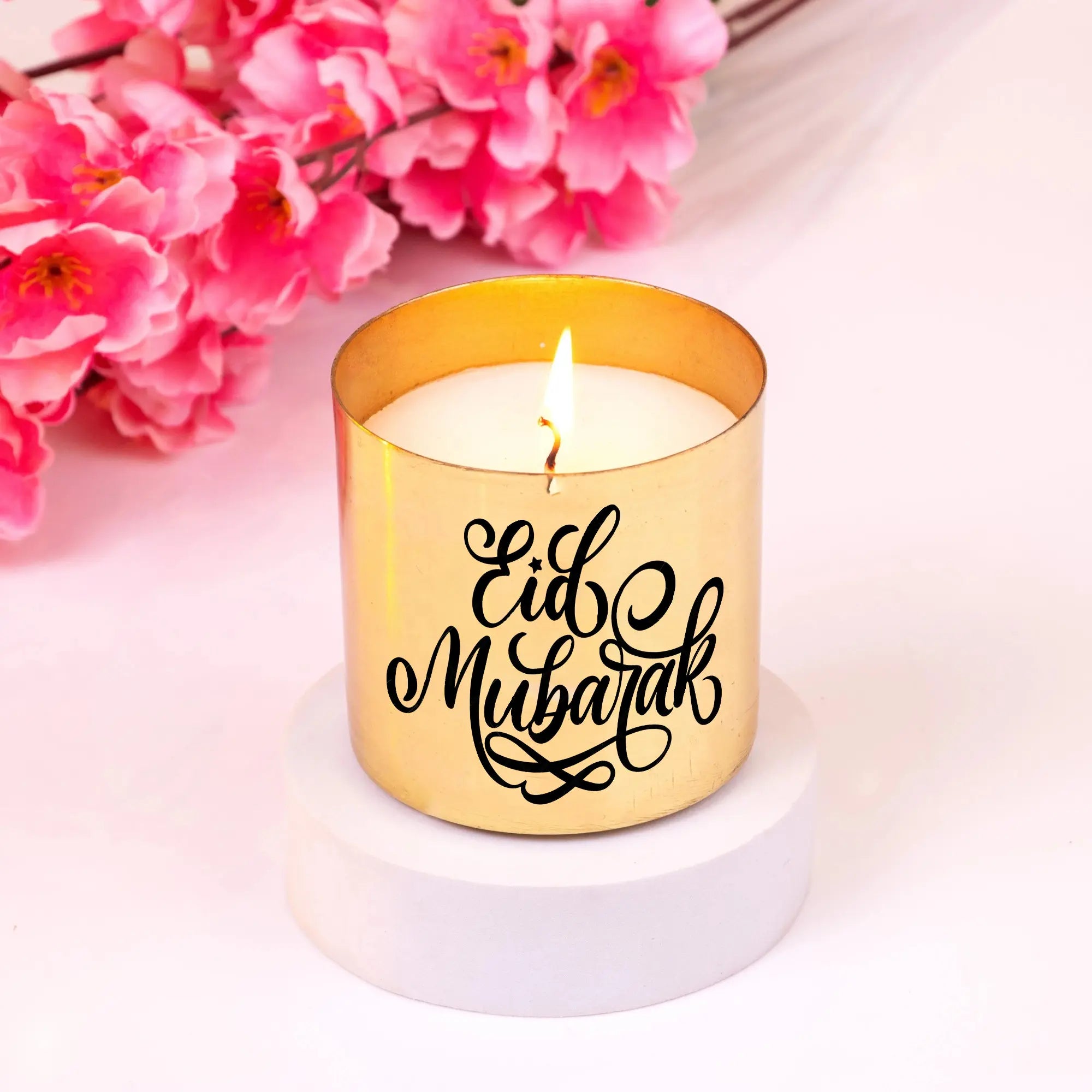 Eid Mubarak Scented Candle in a gold jar with elegant calligraphy, placed on a white base with pink flowers. Luxurious Eid fragrance gift. Main image