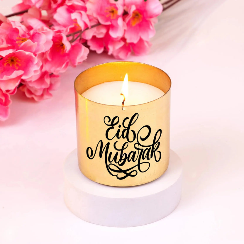 Eid Mubarak Scented Candle in a gold jar with elegant calligraphy, placed on a white base with pink flowers. Luxurious Eid fragrance gift. Main image
