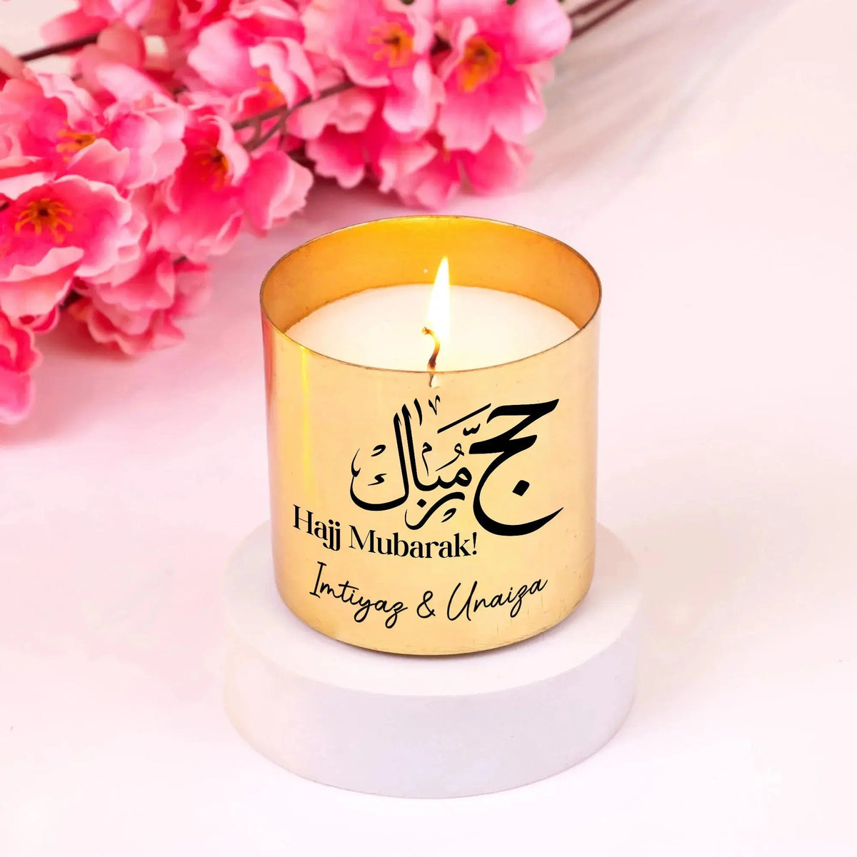 Personalized gold candle from Jannah Journey Hamper - Gift of Faith & Blessings with 'Hajj Mubarak' message, lit and placed on white base.