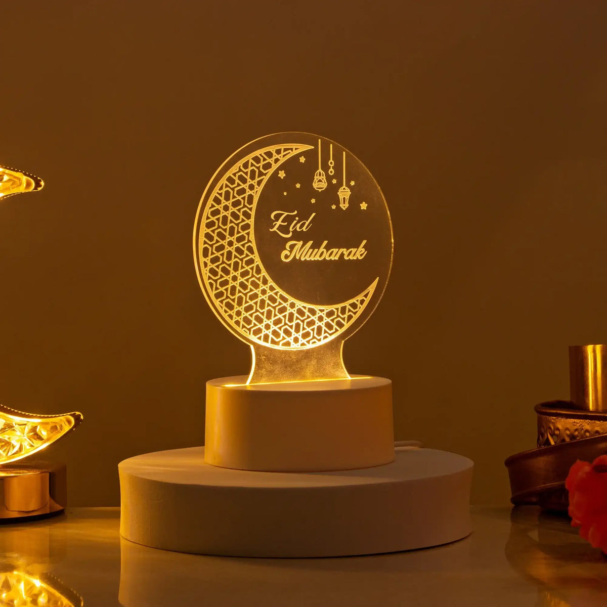 Glowing Eid Mubarak LED acrylic lamp displaying a crescent moon and lantern motif, resting on a white base against a warm, softly lit background.