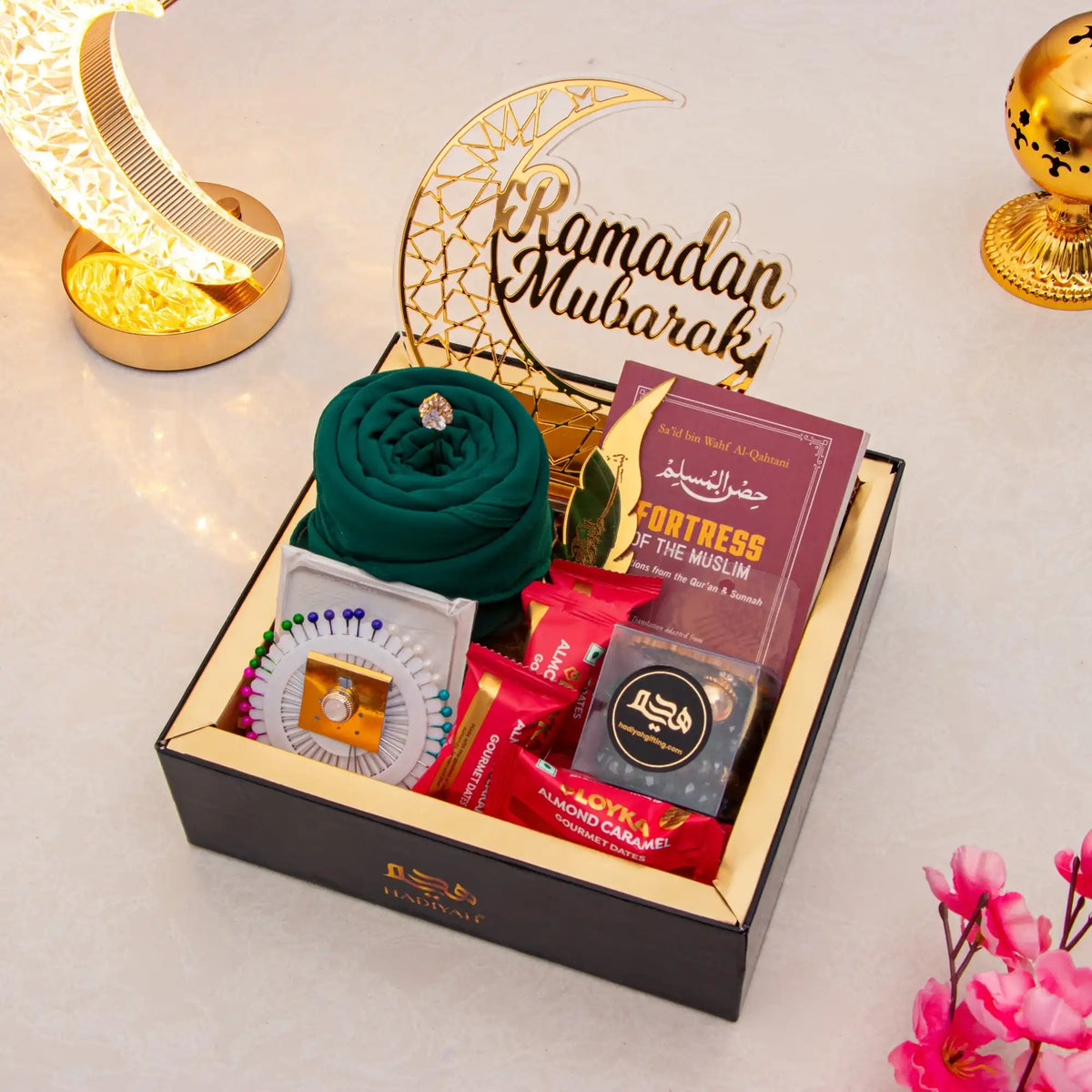 Gift box with Ramadan-themed items including a green headscarf, books, and candies on a decorative background.