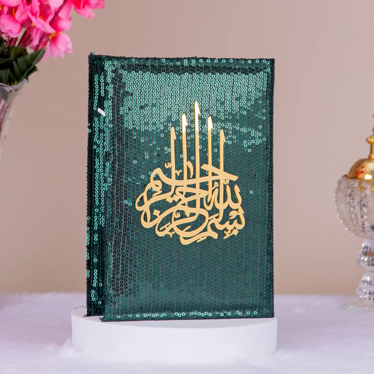 Decorative book with gold text and candle design on a green background, placed on a white surface with pink flowers and a decorative object in the background.