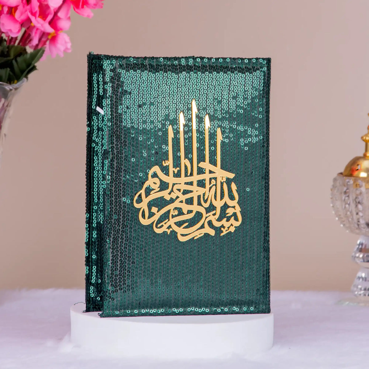 Decorative book with gold text and candle design on a green background, placed on a white surface with pink flowers and a decorative object in the background.