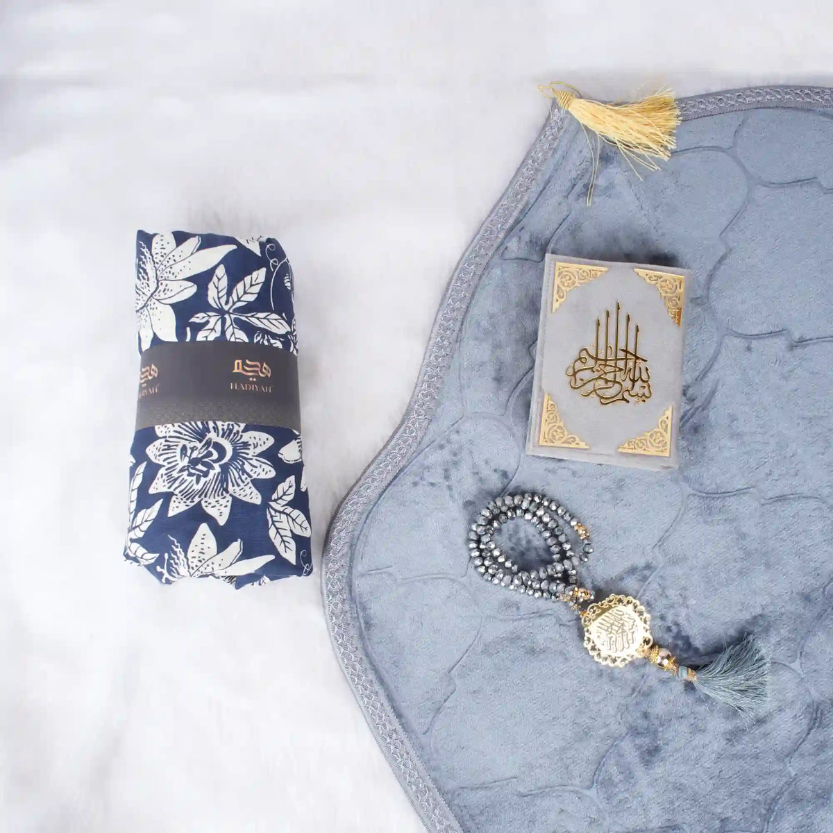 Grey Sajdah Serenity Hamper laid out on a white surface, showcasing a grey velvet janamaz, a mini grey velvet Quran, a grey crystal tasbeeh, and a rolled-up blue and white floral prayer makhana.