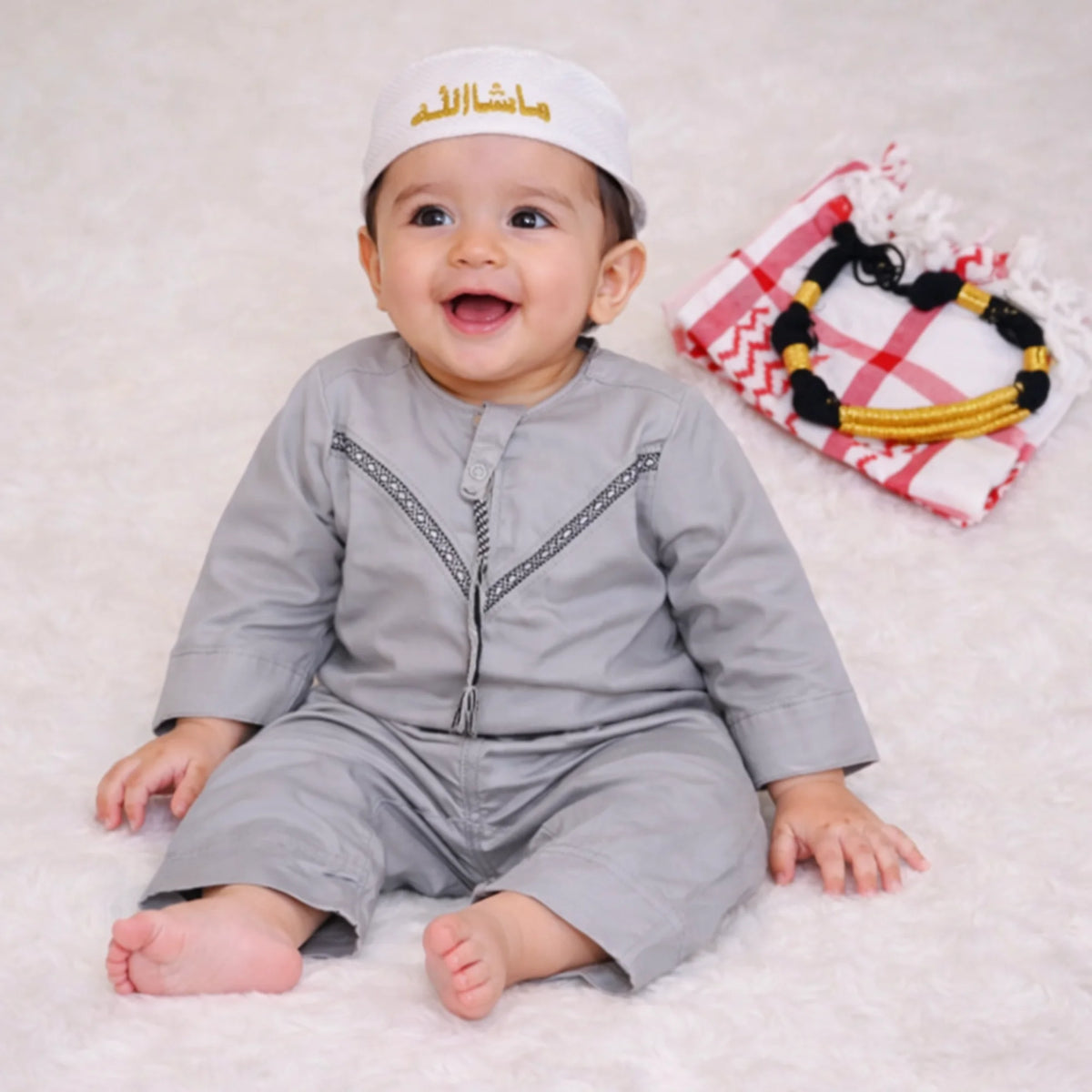 Baby boy in traditional grey thobe with Islamic cap, sitting on soft white surface