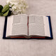 Open book on a light surface with white flowers and green leaves.