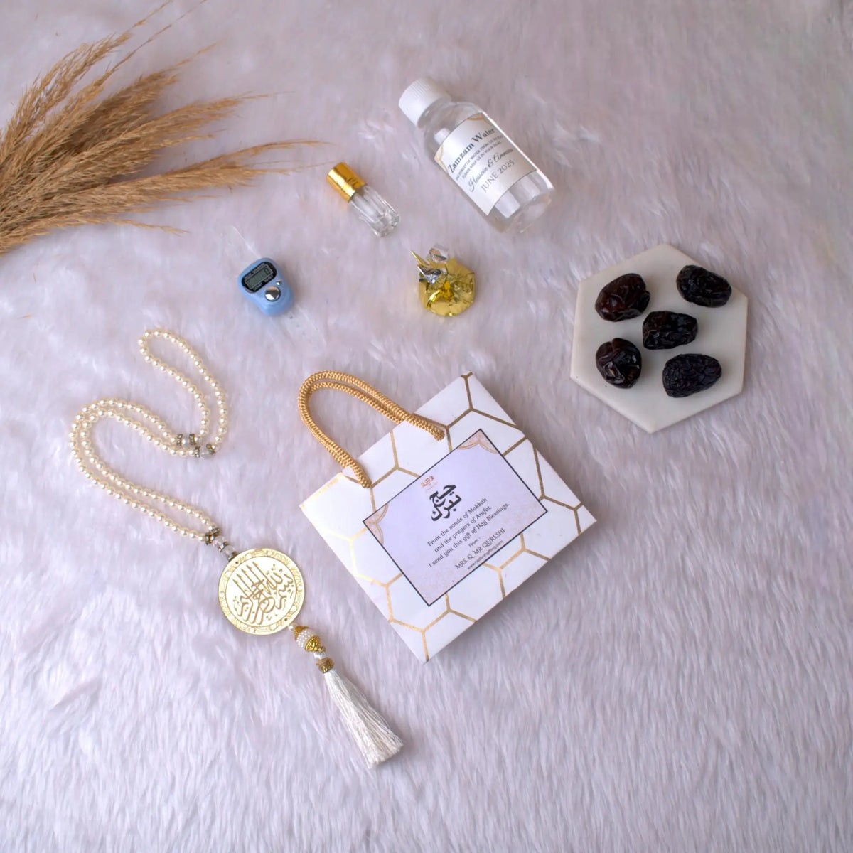 Barakah Hajj Gift Hamper Pack with tasbih, Zamzam water, Ajwa dates, and Islamic accessories on white fur background. Ideal Hajj return gift set.