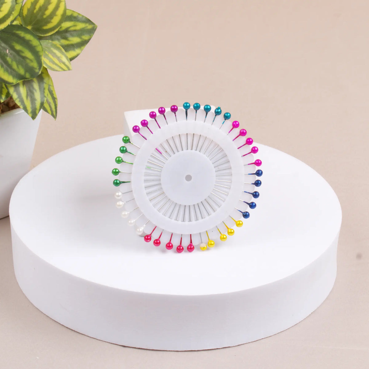 A circular white pin wheel holding numerous straight hijab pins with colorful, pearl-like heads, resting on a white cylindrical display stand next to a small potted plant