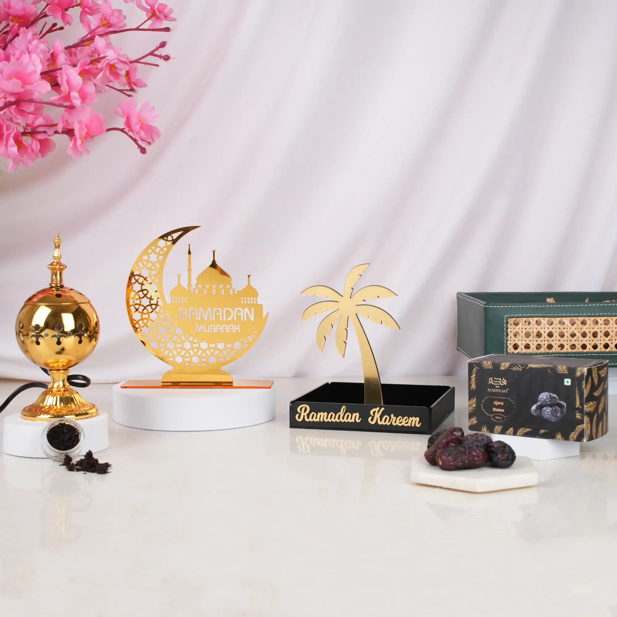 The contents of the Ramadan Ambience Collection displayed individually on a white surface against a white draped background with pink blossoms. It includes a gold electric bakhoor burner with loose bakhoor, a gold Ramadan Mubarak moon and mosque stand on a white pedestal, a black and gold Ramadan Kareem date stand, a box of Ajwa dates with a few loose dates on a coaster, and the green hamper tray in the background. Main image