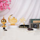 The contents of the Ramadan Ambience Collection displayed individually on a white surface against a white draped background with pink blossoms. It includes a gold electric bakhoor burner with loose bakhoor, a gold Ramadan Mubarak moon and mosque stand on a white pedestal, a black and gold Ramadan Kareem date stand, a box of Ajwa dates with a few loose dates on a coaster, and the green hamper tray in the background.