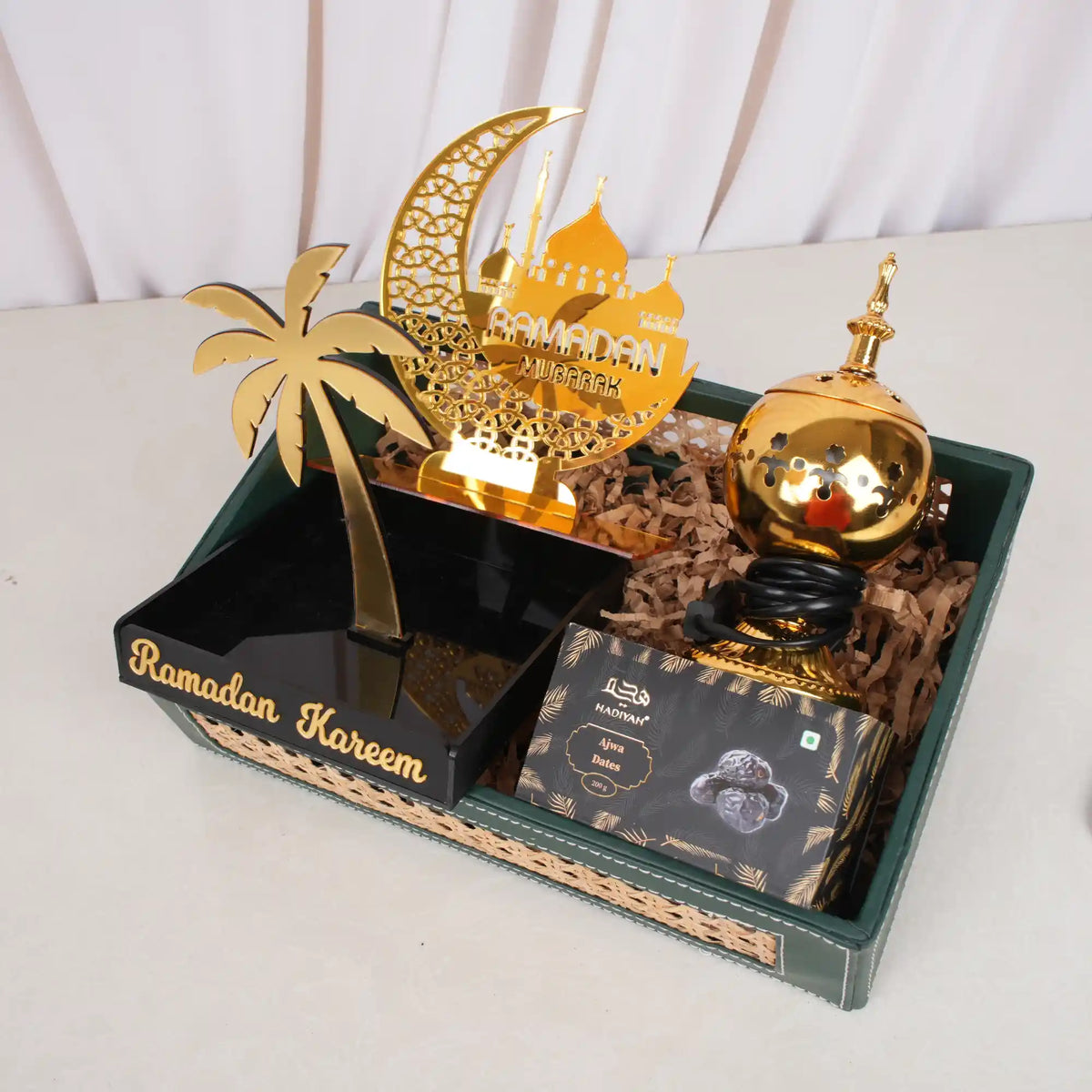 A green woven tray containing the Ramadan Ambience Collection, featuring a gold electric bakhoor burner, a gold acrylic Ramadan Mubarak stand shaped like a crescent moon and mosque, a black acrylic date stand with a gold palm tree and "Ramadan Kareem" text, and a black box of Hadiyah Ajwa Dates nestled on brown shredded paper.