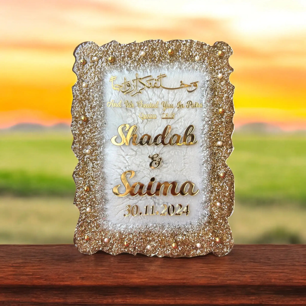 Decorative plaque with 'Shadab & Saima' and date on a wooden surface with a sunset background