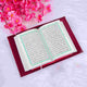 A top-down view of an open Quran with Arabic script resting on a maroon velvet book sleeve. The book is laid on a white faux-fur surface. A large cluster of pink cherry blossoms is visible in the top-left corner, creating a serene and spiritual aesthetic suitable for Ramadan or Eid gifting.
