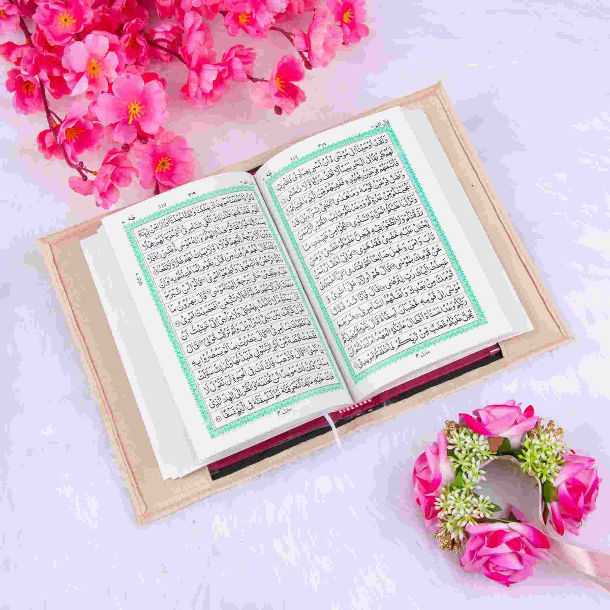 An open Quran with Arabic script is centered on a light beige book sleeve, which rests on a soft, white faux-fur surface. Vibrant pink cherry blossom branches frame the top left corner, while a small floral wreath featuring pink roses and tiny white flowers sits in the bottom right. The image uses a bright, top-down perspective, creating a clean and serene aesthetic often used for Islamic gifting and spiritual content.