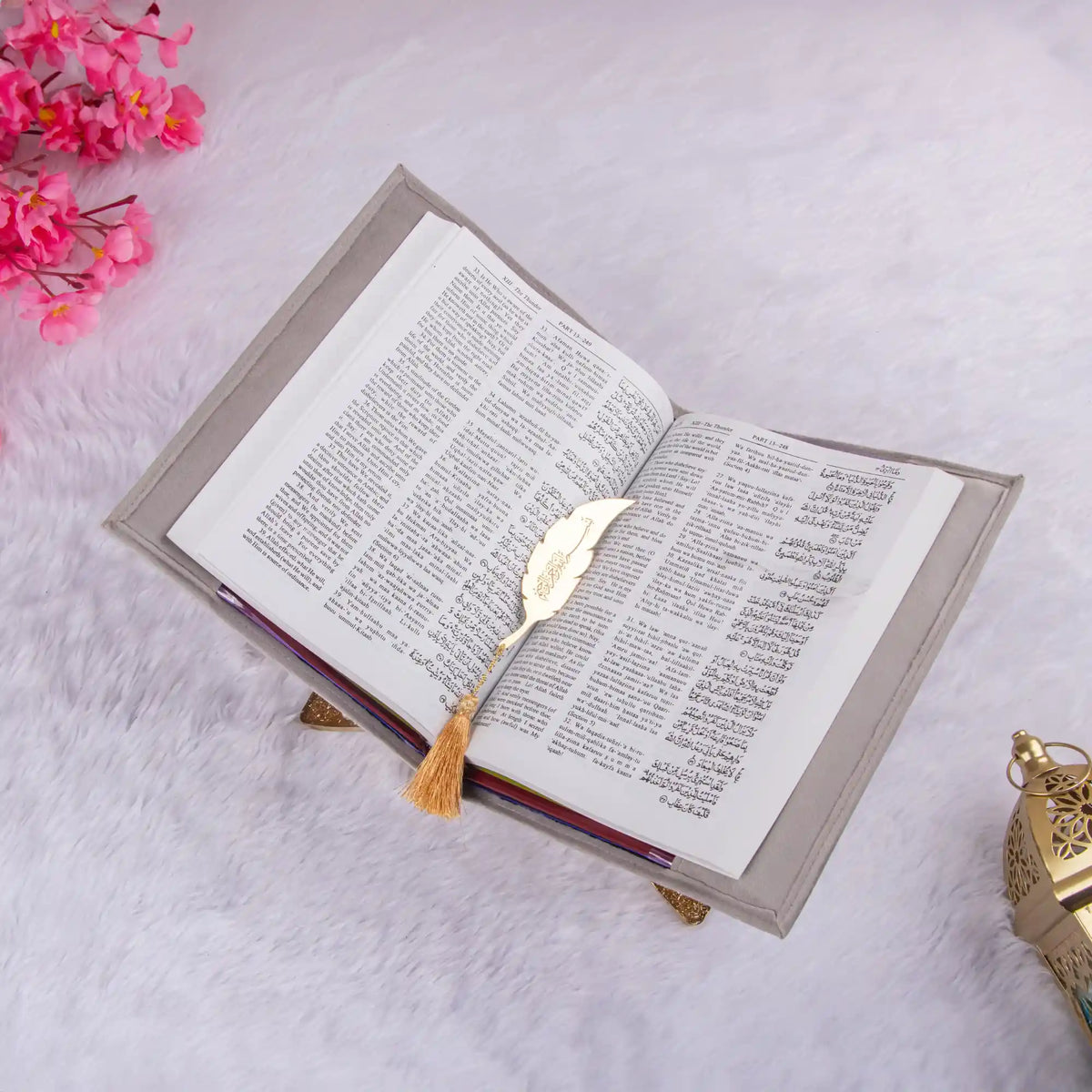 Open book with a decorative gold bookmark on a light pink surface with pink flowers and a small decorative lantern.