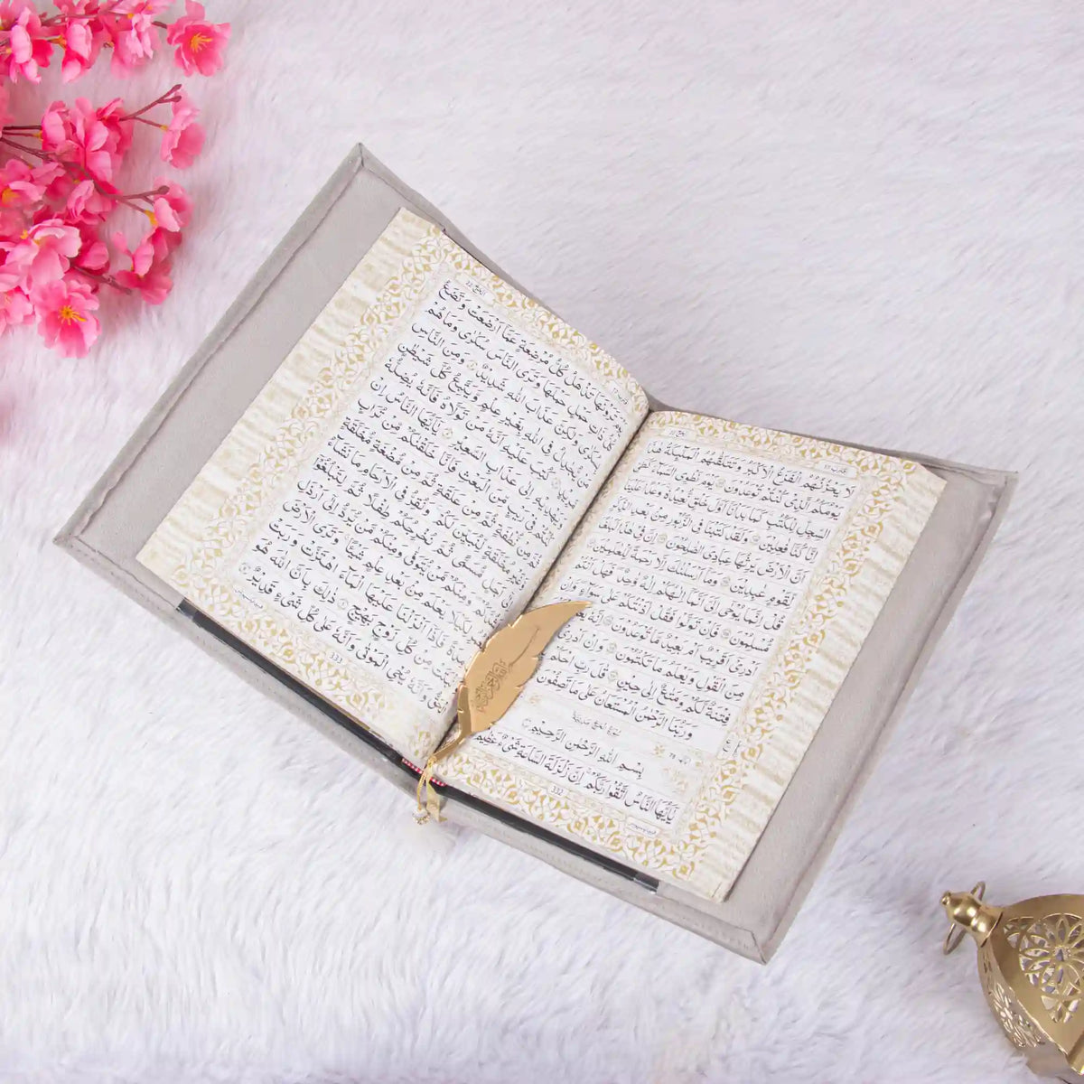 An open Quran with a grey velvet cover laid flat on white faux-fur. A gold feather-shaped metal bookmark rests on the right-hand page, which features Arabic script with a decorative gold border. The image is decorated with pink flowers in the top-left corner and a gold Moroccan-style lantern in the bottom-right.