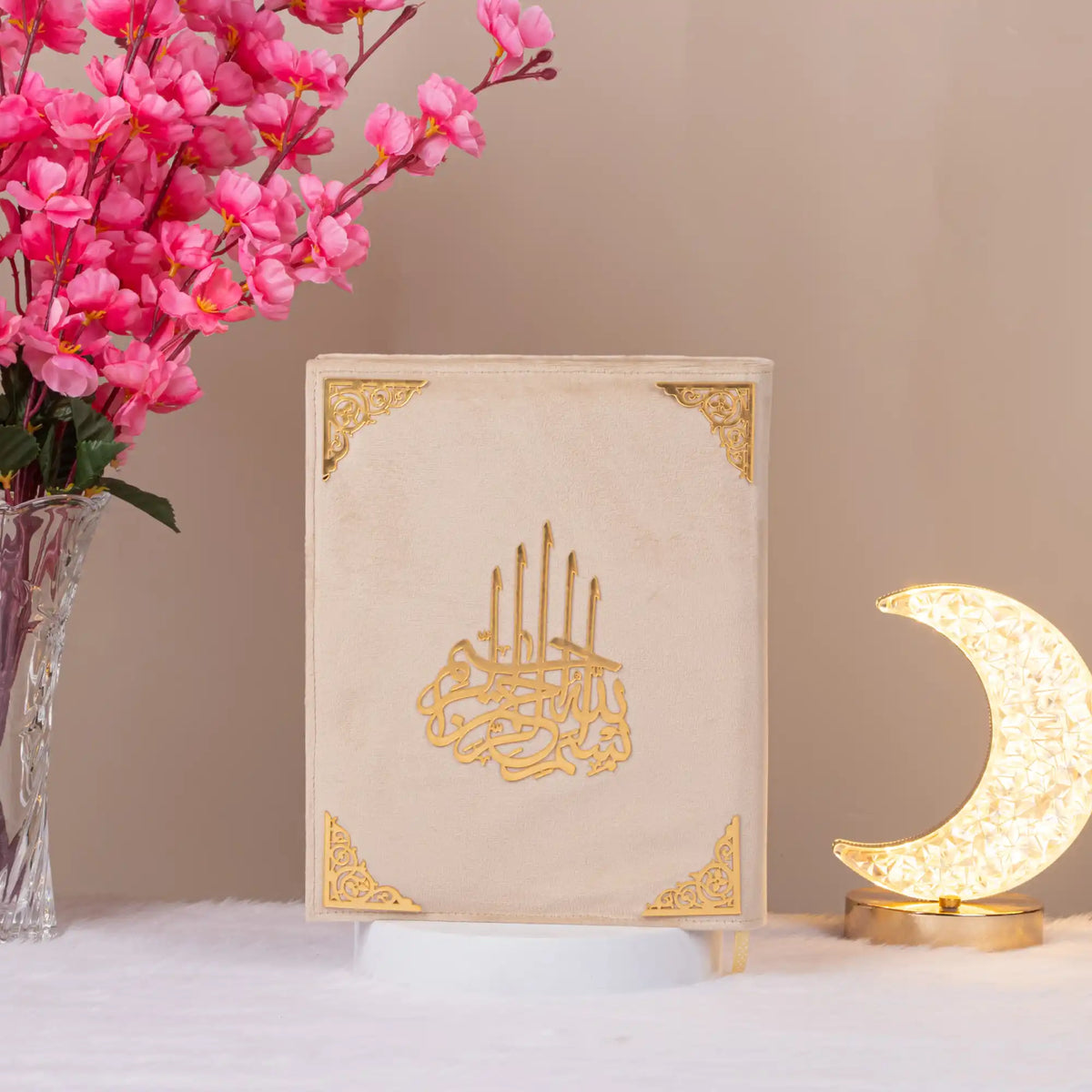 Cream velvet Kaaba Quran with gold accents, standing beside crescent lamp and flowers, perfect for elegant Islamic gifting.