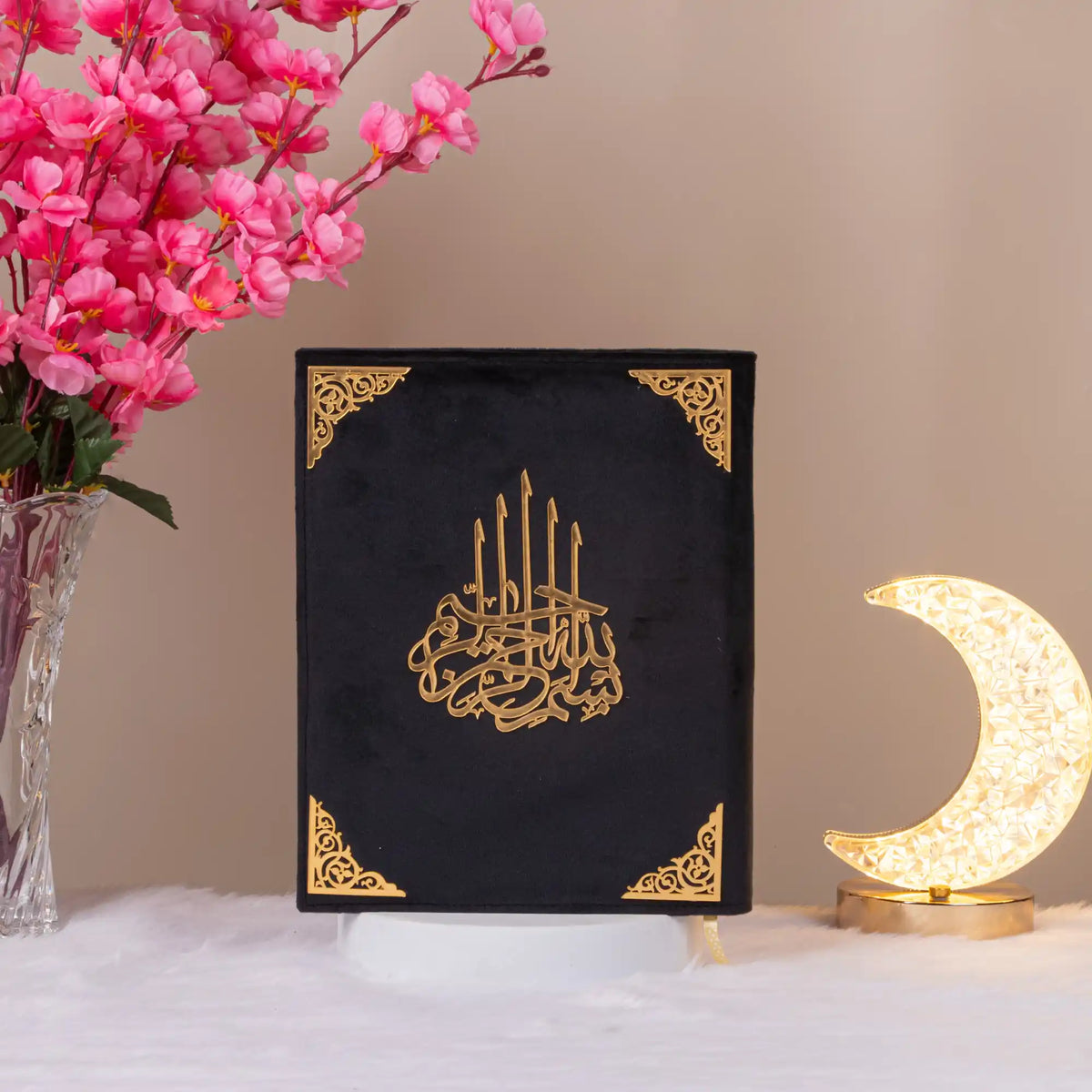 Black velvet Kaaba Quran with gold detailing, displayed upright with floral decor, a refined religious gift for special occasions.