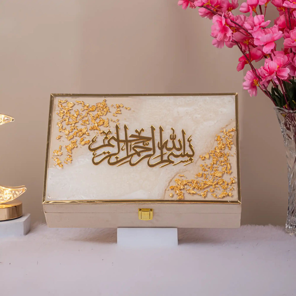 Decorative box with Arabic calligraphy on a stand, surrounded by pink flowers and a small lamp.