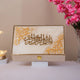 Decorative box with Arabic calligraphy on a stand, surrounded by pink flowers and a small lamp.