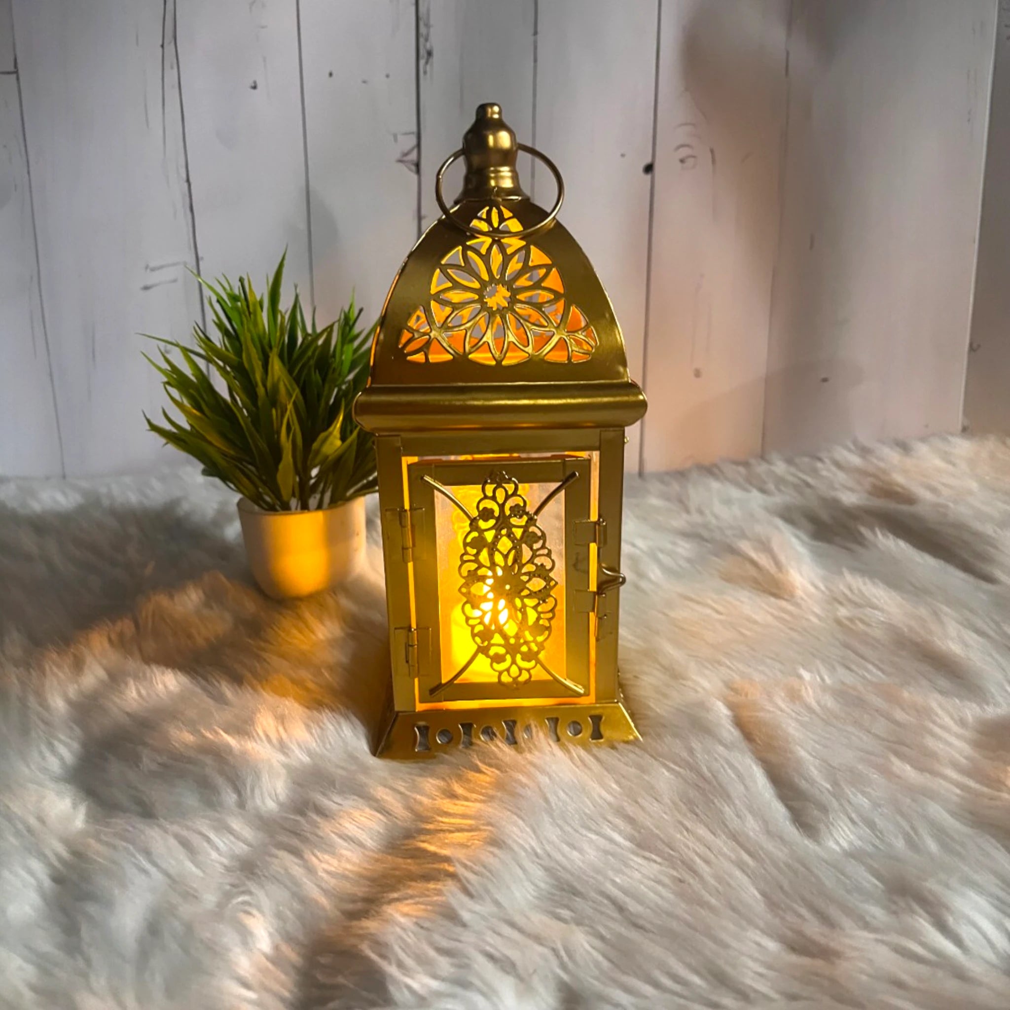 Golden Aura Lantern 9" – Elegant gold finish lantern with intricate floral cutouts, glowing warmly on faux fur with a plant decor backdrop. Main image