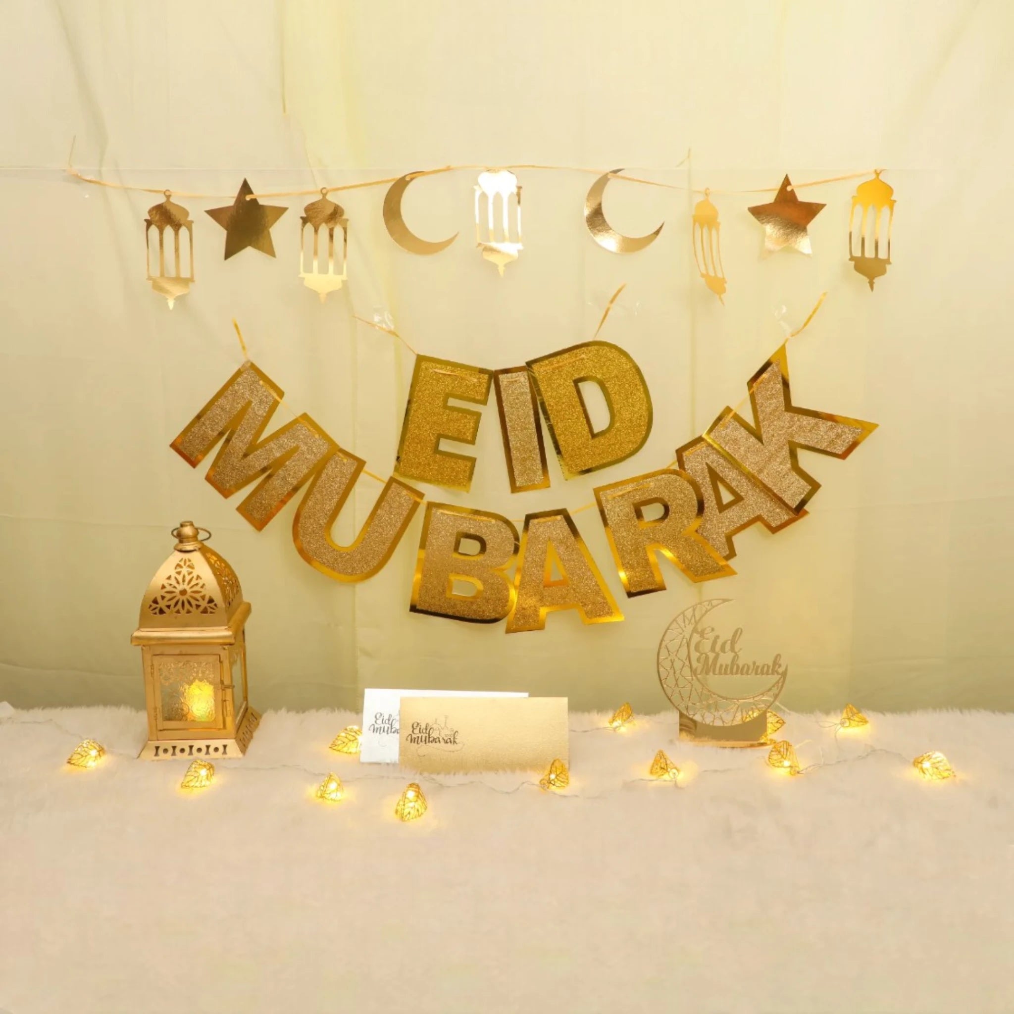 Eid Grand Celebration Decor Set featuring gold Eid Mubarak banner, lantern, moon and star garland, envelopes, and warm string lights. Main image