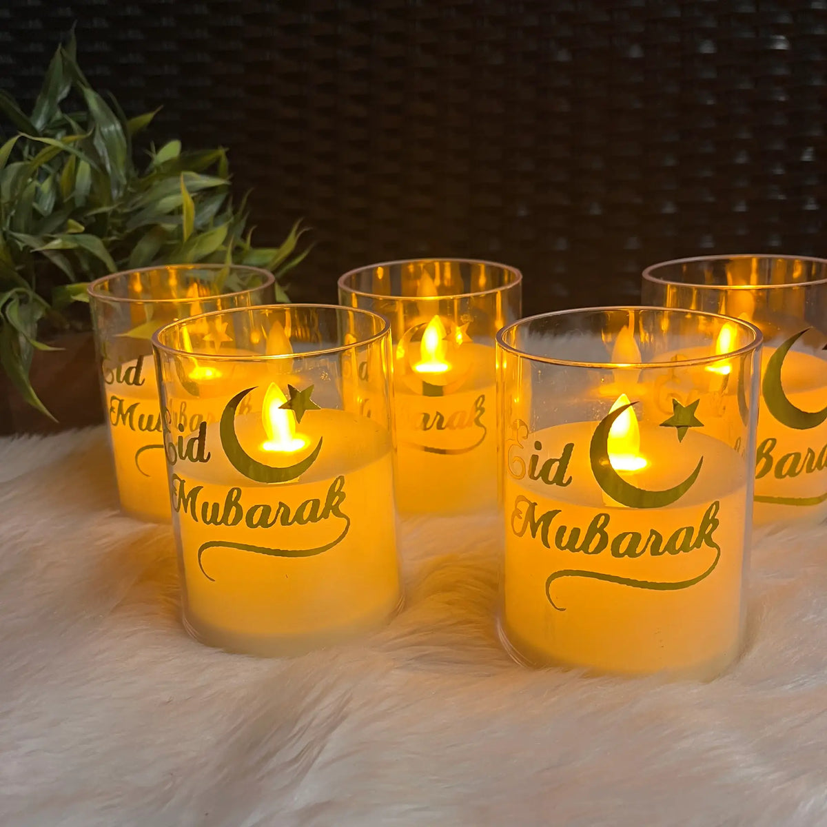 Set of lit candles in glass holders with 'Aid Mubarak' text on a soft surface.