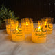 Set of 'Eid Mubarak' candles in glass holders on a soft surface with a plant in the background.