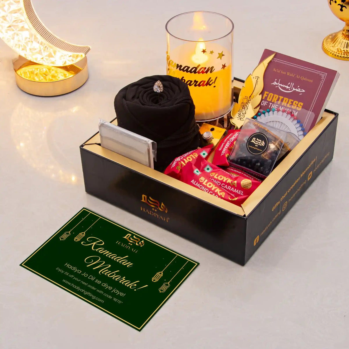 Gift box with Ramadan-themed items including a candle, card, and snacks on a white surface.