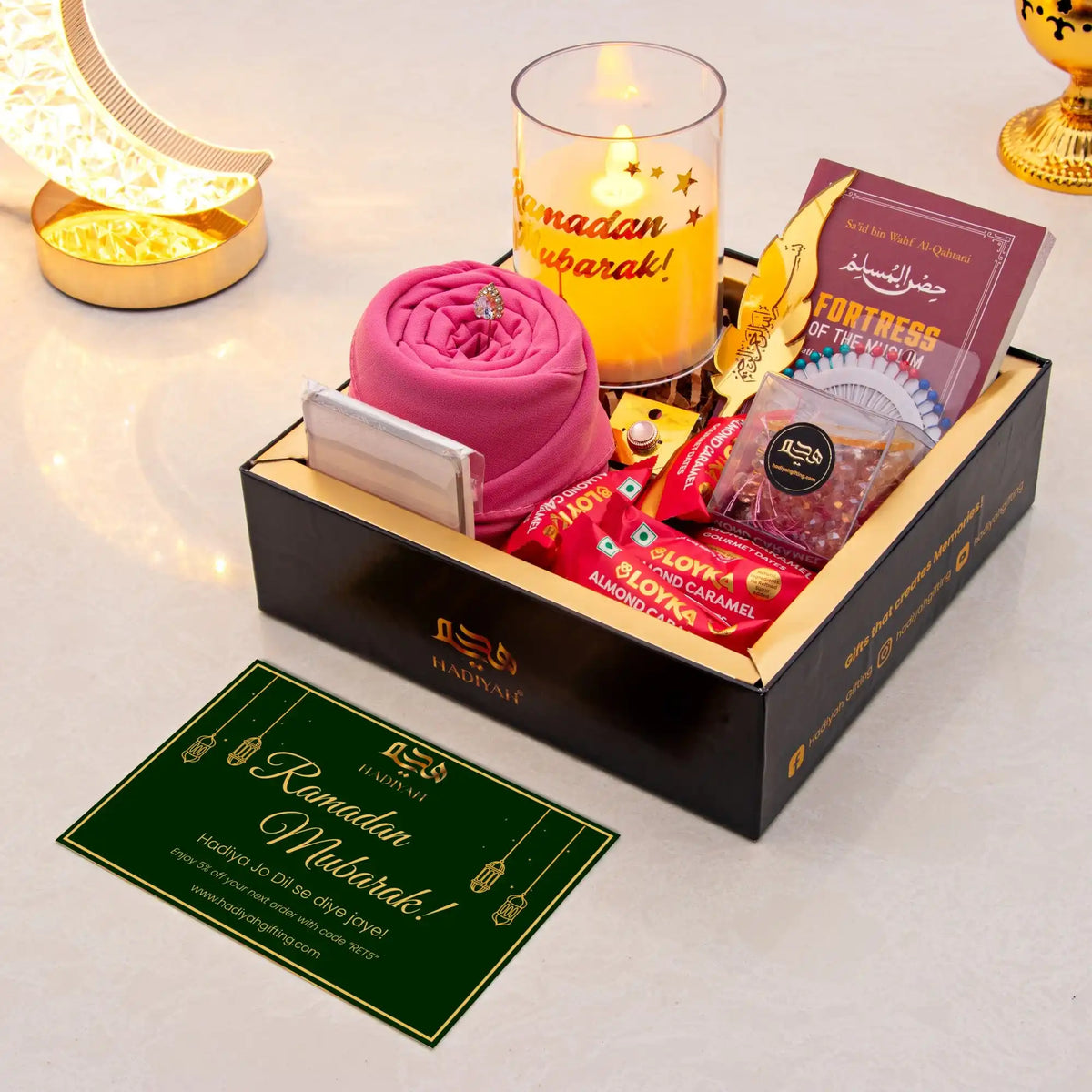 Gift set with pink fabric, candles, and snacks on a white surface with 'Ramadan Mubarak!' card.
