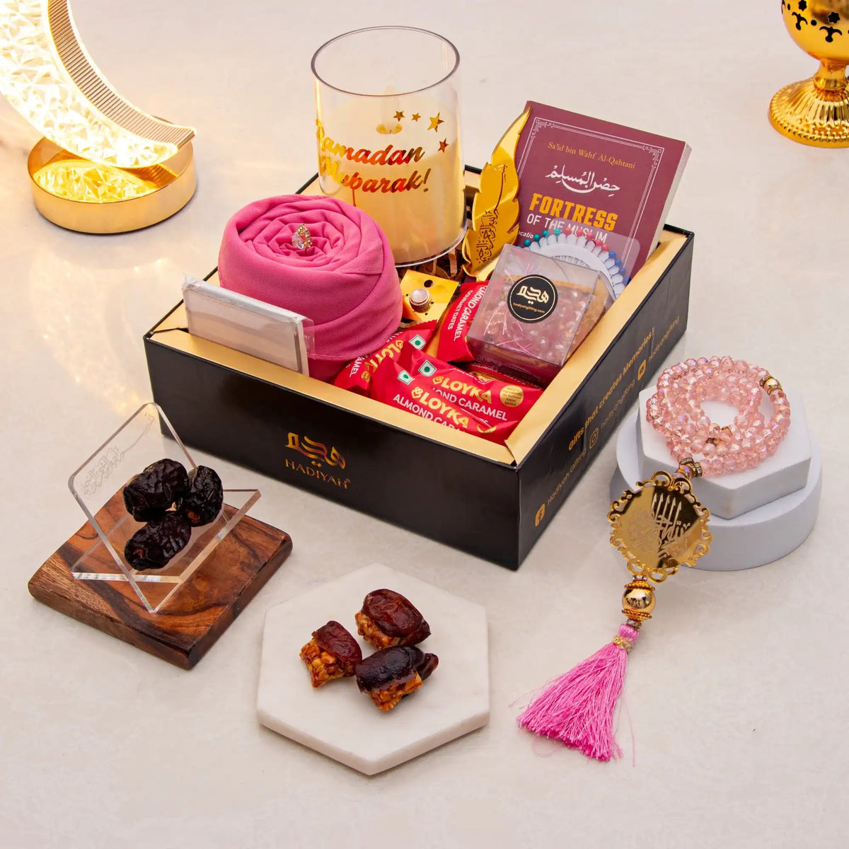 Gift box with snacks, dates, and a rosary on a decorative surface with candles and lights.