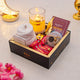 Gift box with snacks, a candle, and a book on a decorative surface with flowers.