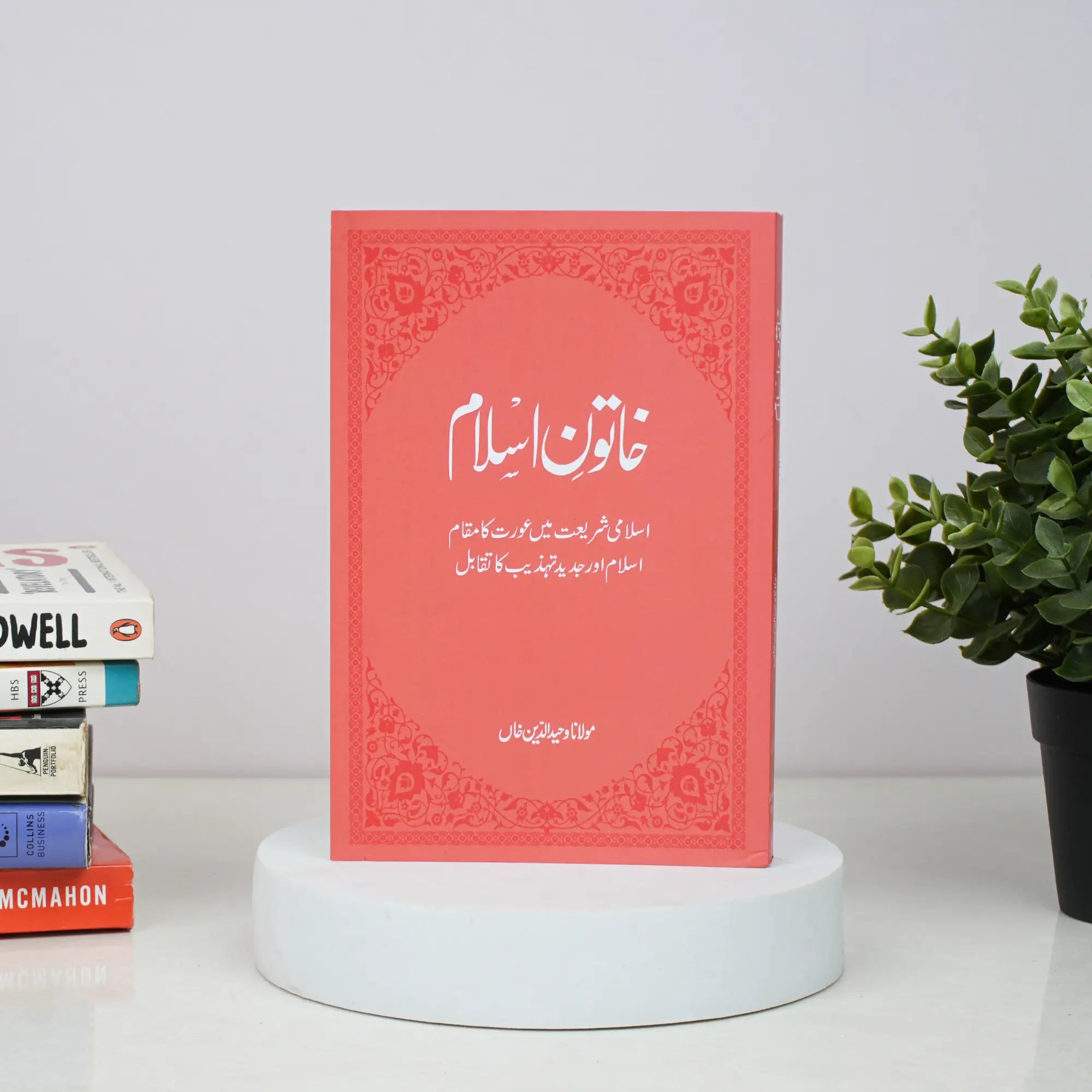 Red book with Arabic text on a white surface with books and a plant in the background