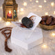 A single dark date resting on a small, clear acrylic holder decorated with festive Ramadan artwork. The holder is displayed on a white hexagonal base against a blurred background featuring a glowing ornate lantern, a beaded rosary, a golden bowl filled with dates, and soft string lights over a white faux-fur surface.
