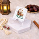 An empty, clear acrylic date stand resting on a white hexagonal block. The stand prominently displays a printed design of a crescent moon and mosque alongside the text "Ramadan Mubarak" in green. The background features a glowing decorative lantern, prayer beads, a cinnamon stick, and a bowl of dates on a soft white rug.