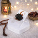 A clear acrylic stand holding a single large date, placed on a white hexagonal base. The stand's backrest features printed Ramadan-themed artwork showing a crescent moon and mosque. The festive background includes a lit brass lantern, wooden prayer beads, a bowl of dates, and warm fairy lights on a fluffy white surface.