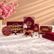 A coordinated set of deep red and gold Islamic wedding stationery pieces arranged on a white faux-fur surface, including a main invitation card, smaller cards, a round name plaque, decorative calligraphy pieces, and a folded red cloth. Pink flowers and warm window-shadow lighting are visible in the background.