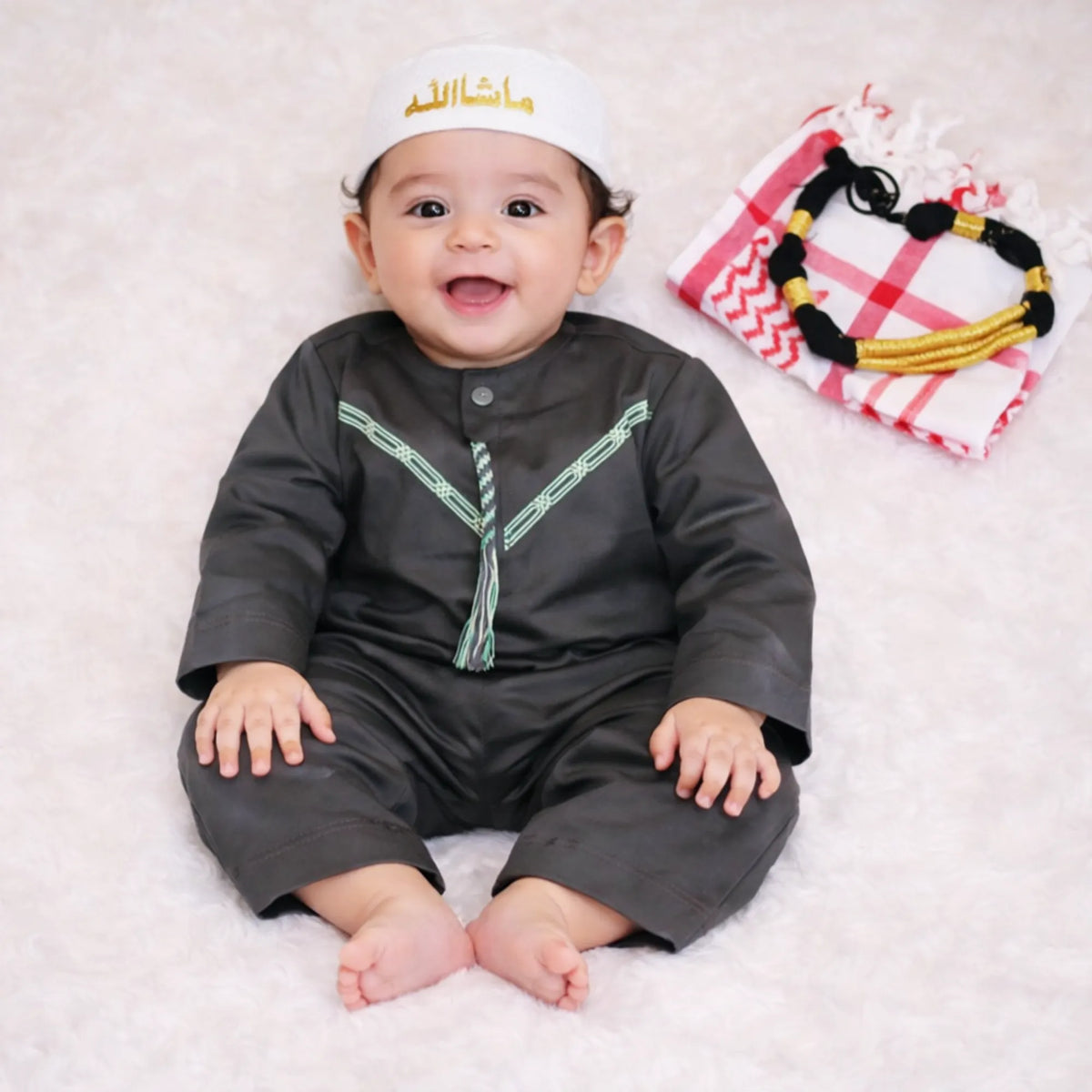 Baby boy in traditional black attire with white embroidery, lying on white plush surface