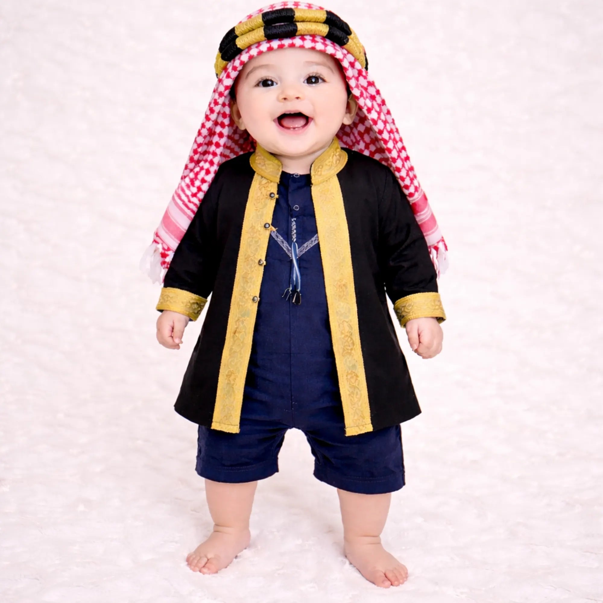 Baby boy in traditional black and gold Islamic outfit with red and white keffiyeh, standing on white textured background Main image