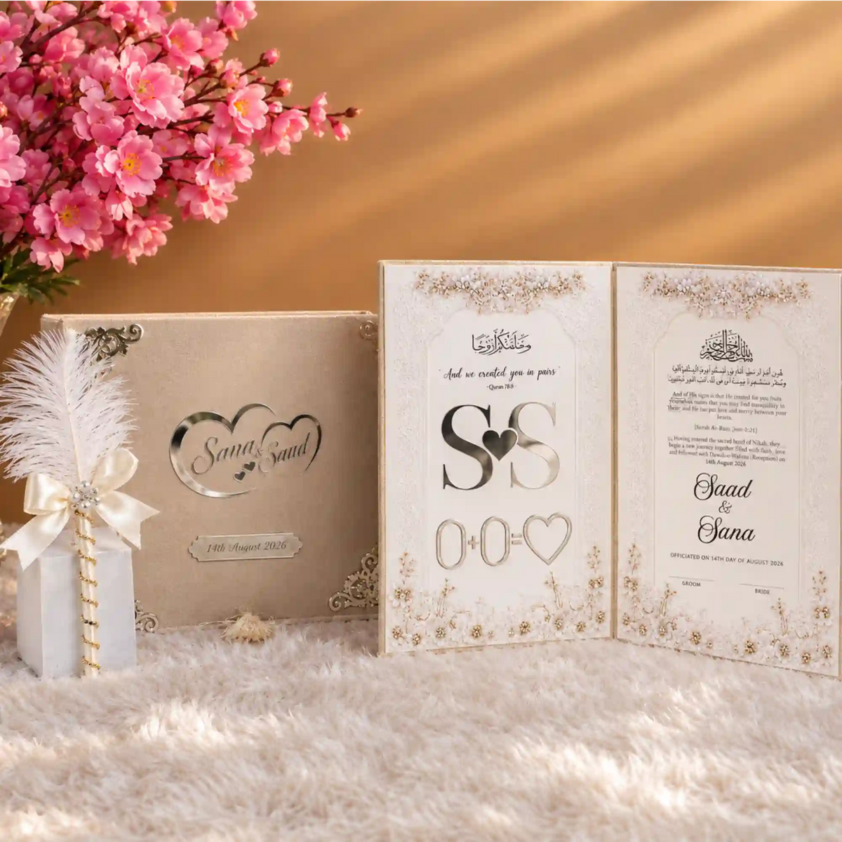 A styled wedding keepsake set featuring a cream-colored textured box with silver corner embellishments and a custom heart design, a matching decorated card with rhinestone corner accents, a white feather pen, and a cream tassel, arranged on a white faux fur surface with pink flowers in the background.