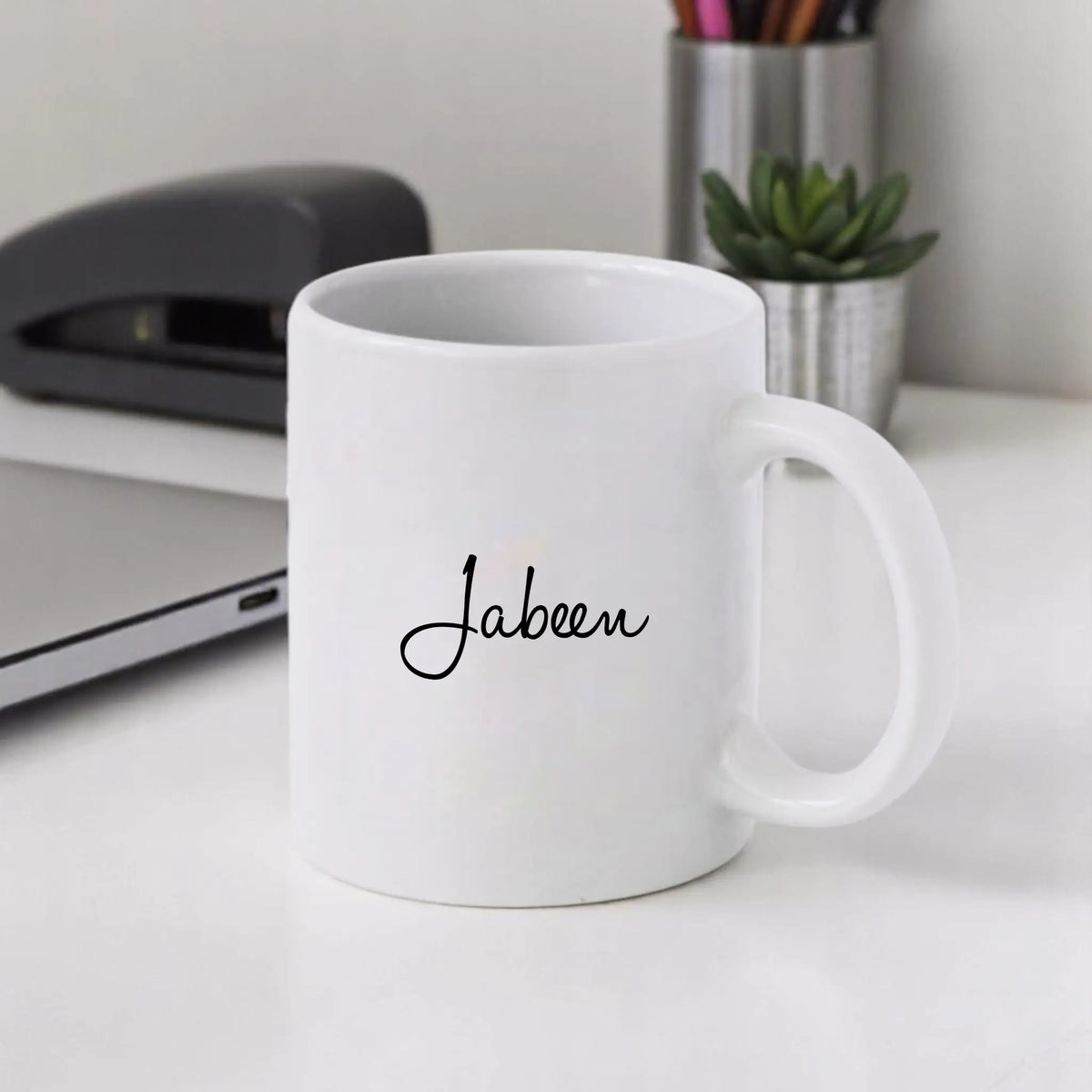 White mug with 'Jabeen' printed on it, placed on a desk with office items in the background.