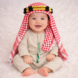 Baby boy in traditional red and white keffiyeh with gold and black headband, sitting on white fluffy surface