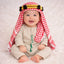 Baby boy in traditional red and white keffiyeh with gold and black headband, sitting on white fluffy surface