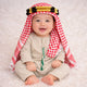 Baby boy in traditional red and white keffiyeh with gold and black headband, sitting on white fluffy surface