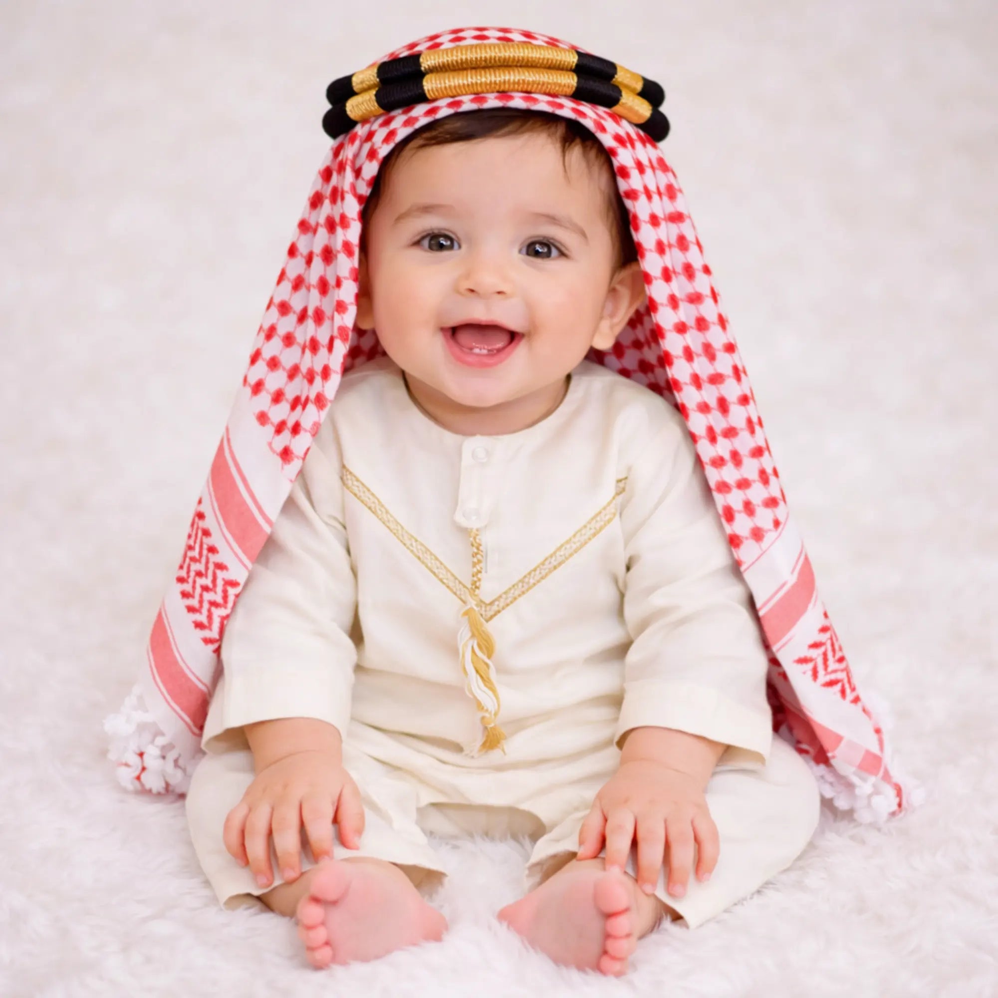Baby boy wearing traditional white thobe with red checkered headscarf Main image
