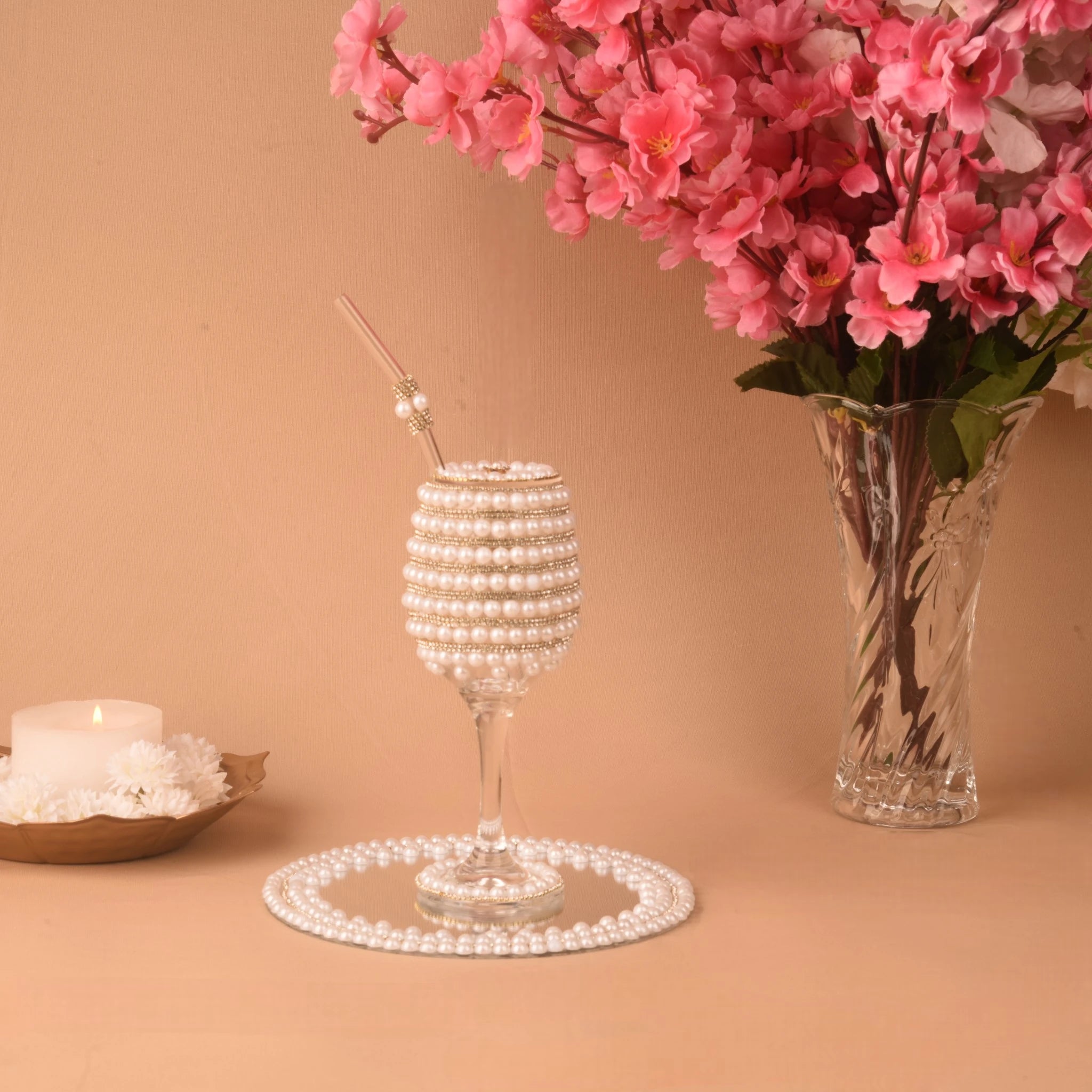 Decorative glass with pearl-like beads, a vase of pink flowers, and a candle on a beige background